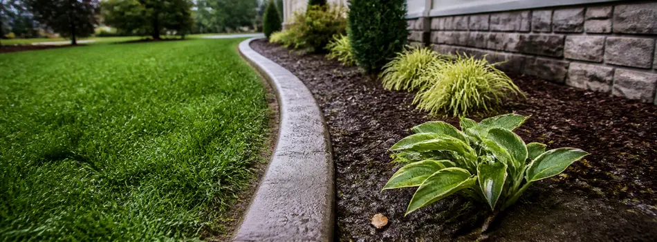 Curved gray concrete curb between a lawn and a mulched bed along a stone house foundation
