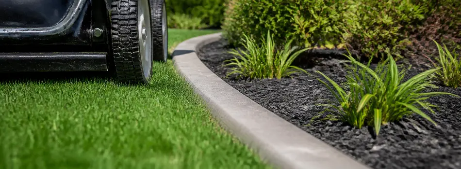 Mower wheel rolling along a smooth concrete curb beside a black-mulch planting bed